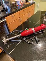 KitchenAid handheld immersion blender red body with attached power cord and detachable blending shaft laid on kitchen counter.
