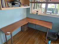L shaped desk with wood finish surface placed against wall beneath window, metal legs visible, curved edges on desk surface.
