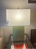 First photo showing one green ceramic table lamp with a rectangular white fabric shade on a glass side table next to a beige armchair.