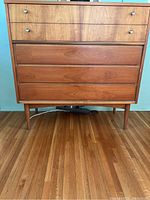 Front view of a wooden dresser with four drawers, two lighter wood drawers on top with metal knobs and three darker wood drawers below, standing on tapered legs on a wooden floor.