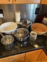 Overview photo showing all kitchen items together on a kitchen counter: a white plastic colander, medium stainless steel pot with glass lid, three nested stainless steel bowls, small stainless steel sauce pot, wooden bowl, and small glass jar.