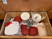Drawer with various kitchen items including several containers with red lids, a manual citrus juicer, small metal strainer, and a clear mixing bowl