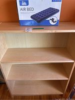 Wooden shelf unit with three shelves, light wood color, with boxed Bedped air bed on top shelf