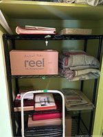 Black metal shelving unit with four open wire grid shelves containing boxes, blankets, and other items. In front of shelf is a white metal step stool with black plastic steps.