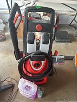 Front view of the red and black Sun Joe electric pressure washer with hose coiled at the front and cleaning product bottles on the floor below.