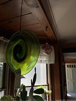 Green hanging glass disc with black spiral swirl pattern photographed suspended indoors against a window background.