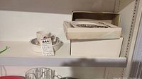 Child's dishes with cup and bowl and boxed silver plated flatware on white shelf