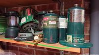 Shelf view showing green Coleman propane lantern next to multiple propane fuel canisters, replacement lantern globes in packaging, and a battery-operated camping light.