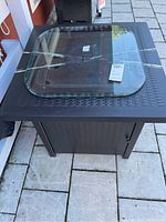 View of square outdoor fire pit with woven rattan-style metal base and clear tempered glass top with rounded corners placed on top