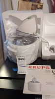 White and silver Krups ice cream maker with 1.5 quart bowl, wrapped in plastic, including motor unit and power cord, placed in original Styrofoam packaging. Manuals also present.