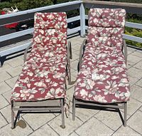 Two outdoor metal patio lounge chairs with puffy floral cushions. One cushion is more weathered with faded colors and some dirt marks. Cushions have a red background with white floral pattern.