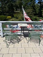 Two green metal outdoor rocking chairs with leaf decorative elements, placed outside on a tiled patio against a white railing with an American flag in background.