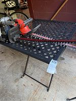 Table with lattice metal top and foldable legs in a garage setting. Various tools placed on top but not included.