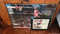 Wide shot of framed Michael Jordan dunk poster alongside framed Joe Namath Super Bowl III collectible photo and article resting on wooden floor against wall