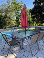 Full view of the rectangular metal patio table with glass top, surrounded by six chairs and closed red umbrella through the table center.