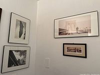 Four black and white architectural and travel-themed prints framed and hung on a white corner wall. Prints include images of a pedestrian on a bridge, close-up of sculpture, and building elements. Frames are black with some wear.