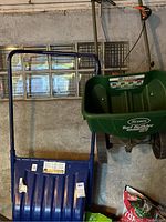 Blue plastic wheelbarrow with metal frame and a green Scotts Turf Builder seed spreader next to it, both stored in a garage with concrete block walls.
