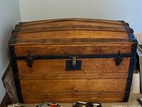 Front view of solid wood trunk showing curved lid, metal corner protectors and black latch hardware, with rustic aged wood finish.