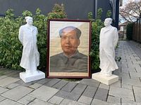 Front view of two white statues and framed portrait of Chairman Mao on outdoor pavement with greenery background.