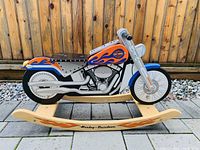 Full side view of wooden rocking horse shaped like a Harley-Davidson motorcycle on a stone tile surface with wooden fence background.