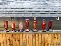 Seven glass hummingbird feeders lined on an outdoor wooden ledge under a roofline, showing full lot in natural light.