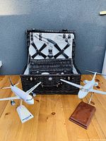 Photograph showing the black woven picnic basket with interior setup of plates and utensils alongside two airplane models on stands.