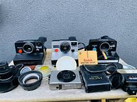 Photo showing three Polaroid cameras, Canon ELPH digital camera, several lenses and round camera accessories on a wooden surface against a gray wall.