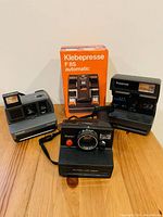 Front view of three Polaroid cameras and Klebepresse F8S automatic box on wooden surface