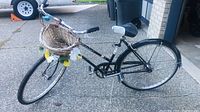 Side angle view of black Schwinn female adult bike showing wicker basket decorated with flowers on the front, frame design, and rear wheel.