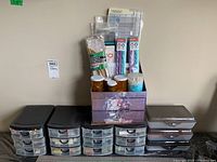 Photo showing multiple plastic storage drawers with black lids and a floral decorative two-drawer box on top containing various small containers and art supplies.