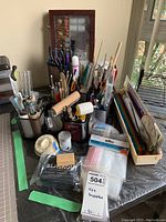 Wide view of various art supplies including paintbrushes, rollers, pliers, dremel bits, and bags organized in mesh holders on a table.