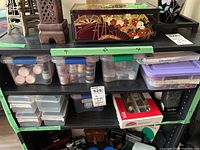 Shelf with multiple plastic boxes containing art supplies including paints, glitter, sewing threads, Dremel bits, and a decorative box on top