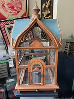 Front view of the bird cage showing the wood frame, metal wire bars, arched door and blue peaked roof with finial.