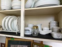 Shelves filled with multiple white plates, bowls, soup spoons, ramekins, and other assorted white ceramic kitchenware.