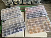 Top view of four plastic compartment storage boxes filled with assorted metal rings (jump rings and split rings) in silver, gold, rose gold, black, purple, and other finishes, organized by size