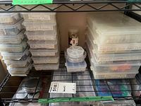 Shelf with multiple plastic organizing containers stocked with labeled compartments holding crafting jewelry findings such as brooch pins, closed rings, and metal components.