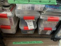 Stacked clear plastic containers with red handles labeled as crafting supplies on a green tape.