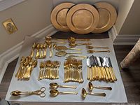 Full set of gold colored forks, spoons, knives, and serving utensils laid out on fabric, with large round gold-colored plates stacked behind.