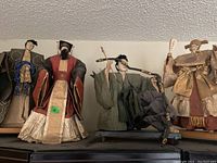 Four Oriental style figurines displayed in a row on a shelf, showing full bodies and diverse costumes.