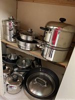 Stainless steel cookware collection featuring double boiler, stock pot, frying pans, saucepans with lids, and black-handled utensils.