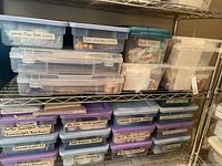 Photo showing a metal shelf with multiple labeled plastic storage boxes containing various crafting supplies including leaves, moss, beads, stems, fruit, vegetables, sewing notions, ribbons, bindings, etc.