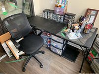 Photo showing black mesh office chair alongside folding table desk with various small storage drawers underneath and many art supplies on top of desk.
