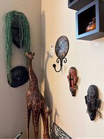 Wall corner showing metal art hook with circular decorative plate and two candle holders, three wood masks mounted on the wall, and part of two giraffe figures in front (not included).