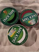 Three Duck Tape rolls in red, white, and black with green branded lids.