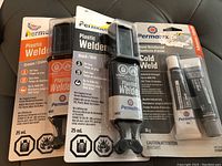 Photo showing three Permatex adhesive products: two Plastic Welders (cream and black) and one Steel Reinforced Cold Weld epoxy epoxy adhesive.