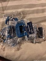 Ten blue metal 1 inch C-clamps, each individually packaged in clear plastic, on a fabric surface.