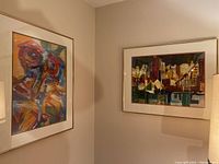 Two framed original paintings by Pablo Schoenefuhs hung on a beige wall. One features an abstract composition of bold and colorful brush strokes, the other a stylized cityscape.