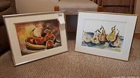 Two framed original artworks by Pablo Schoenefuhs displayed together on carpet. One is a still life with banana, apples, and a pottery pitcher, the other features pears.