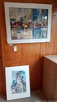Framed abstract cityscape painting by Pablo Schoenefuhs hanging on a wooden wall with a smaller framed painting below on the floor.