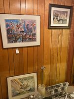 Three framed artworks displayed on wood-paneled wall. One large colorful abstract scene and two smaller paintings including a marina and a sailboat scene.